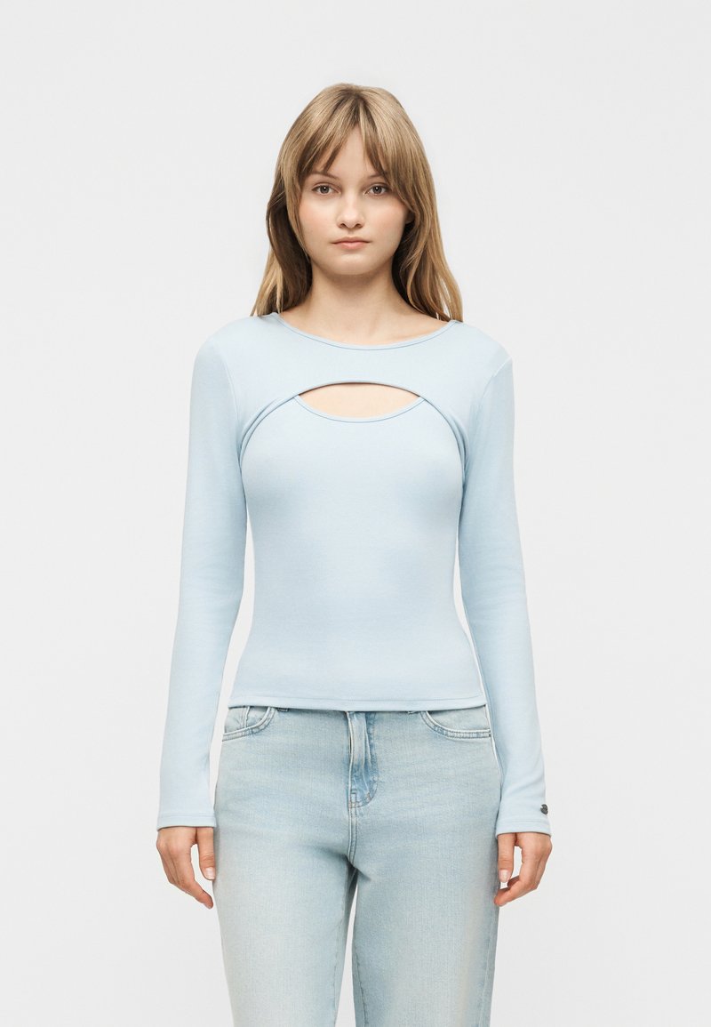 Jauna sieviete valkā gaiši zilu garām piedurknēm topu ar izgriezumu krūtīs un gaiši mazgātas džinsas, stāvot pret vienkrāsainu fonu.