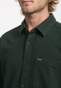 Chemise en flanelle verte avec une fermeture à boutons à l'avant, boutons noirs, et une poche poitrine arborant une étiquette logo. Texture douce et design de col classique.