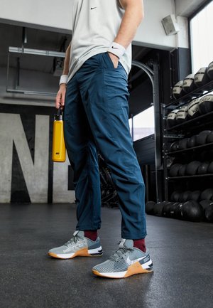Persona que lleva pantalones azules de Nike, zapatillas grises y naranjas, muñequeras blancas de Nike y sostiene una botella de agua amarilla de Nike en un gimnasio.