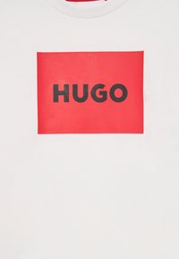 Bílý tričko s červeným obdélníkovým potiskem, na kterém je slovo "HUGO" v tučných velkých černých písmenech. Kulatý výstřih a bavlněný materiál.