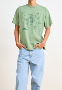 Camiseta de algodón verde oliva con un diseño gráfico de contorno y texto, combinada con jeans de mezclilla azul claro. Corte relajado y mangas cortas.