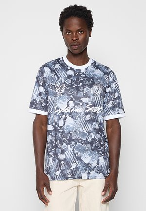 Unge mand iført en sort og hvid T-shirt med grafisk print og hvide kanter, beige bukser, står mod en ensfarvet lys baggrund.