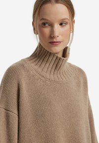 Beżowy sweter dzianinowy z wysokim prążkowanym kołnierzem, szerokimi rękawami i fakturowaną powierzchnią. Charakteryzuje się luźnym krojem i minimalną ilością szwów.