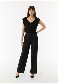 Schwarze Bluse mit kurzen Ärmeln und V-Ausschnitt, kombiniert mit hoch taillierten schwarzen weit geschnittenen Hosen und schwarzen spitz zulaufenden Absatzschuhen.