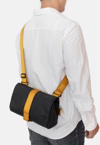 Borsa a spalla in tessuto nero con una tracolla gialla, forma rettangolare e chiusura a patta. Include una tracolla regolabile e dettagli laterali.