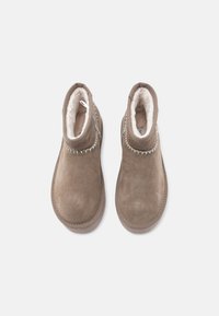 Botas de tornozelo em camurça castanha com um forro branco suave. Apresenta detalhes em costura ao longo do colarinho e um laço para facilitar o calçar.