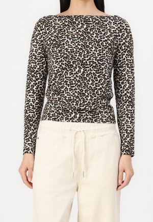 Vrouw die een lange mouwen top met luipaardprint draagt, ingestopt in roomkleurige broek met trekkoord, staand tegen een effen achtergrond.
