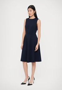 Granatowa sukienka bez rękawów z dopasowaną górą i rozkloszowanym dołem; gładka tkanina o matowym wykończeniu; modelka ma na sobie czarno-białe buty na obcasie w szpic.