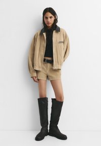 Beige corduroy-takki mustalla kauluksella, yhdistettynä samansävyisiin shortseihin ja mustiin polviin asti ulottuviin saappaisiin, joissa on sileä pinta ja metalliset yksityiskohdat.