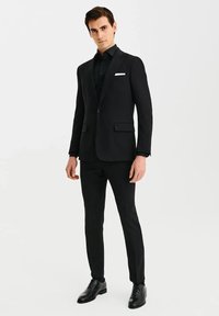 Czarny garnitur z prostymi spodniami, jednorzędową marynarką, klapami w kształcie notcha oraz białą poszetką. Błyszczące czarne buty na sznurówki.