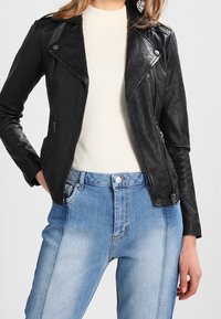 Černá kožená motorkářská bunda se stříbrným kováním, zipovými kapsami a přiléhavým střihem, nošena přes krémový žebrovaný svetr a světle modré rifle.