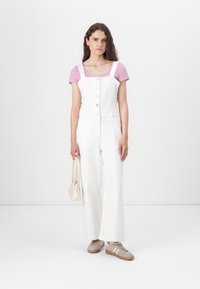 Overoles de denim blancos con grandes botones frontales, corte de pierna ancha, combinados con una camiseta de manga corta rosa y zapatillas beige. Lleva un bolso pequeño.