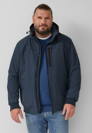 Marineblauwe softshell jas met een afneembare capuchon, een ritszak aan de voorkant en ribgebreide manchetten. Draagt comfortabel over een blauwe trui en lichte jeans.