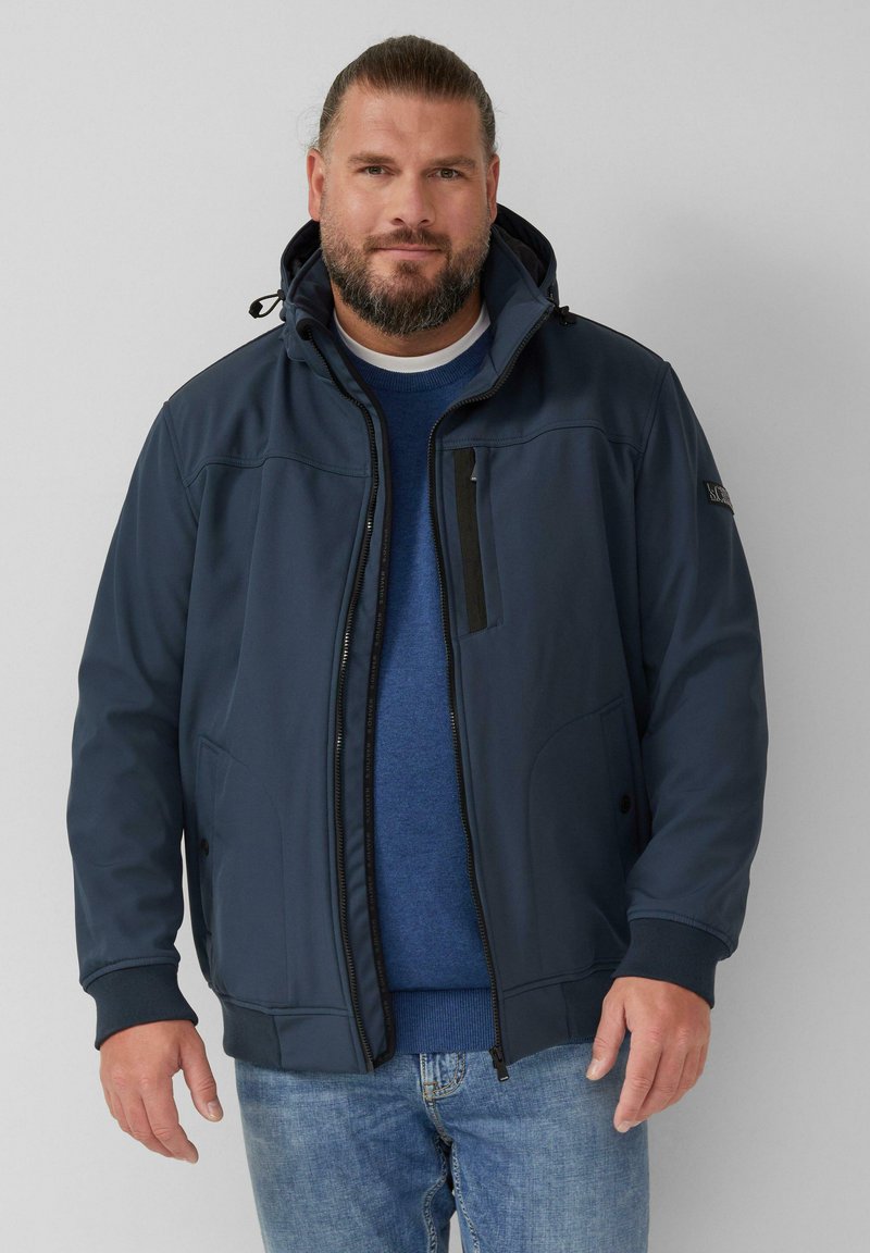 Veste softshell bleu marine avec capuche amovible, poche avant zippée et poignets côtelés. Portée par-dessus un pull bleu et un jean clair.