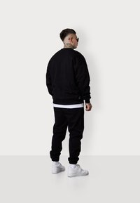 Sweatshirt noir et joggings en mélange de coton, avec une coupe décontractée. T-shirt blanc visible, associé à des baskets blanches.