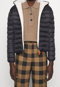 Doudoune noire avec un extérieur texturé, une capuche doublée en polaire et une fermeture éclair. Portée sur un polo beige et un pantalon à carreaux.