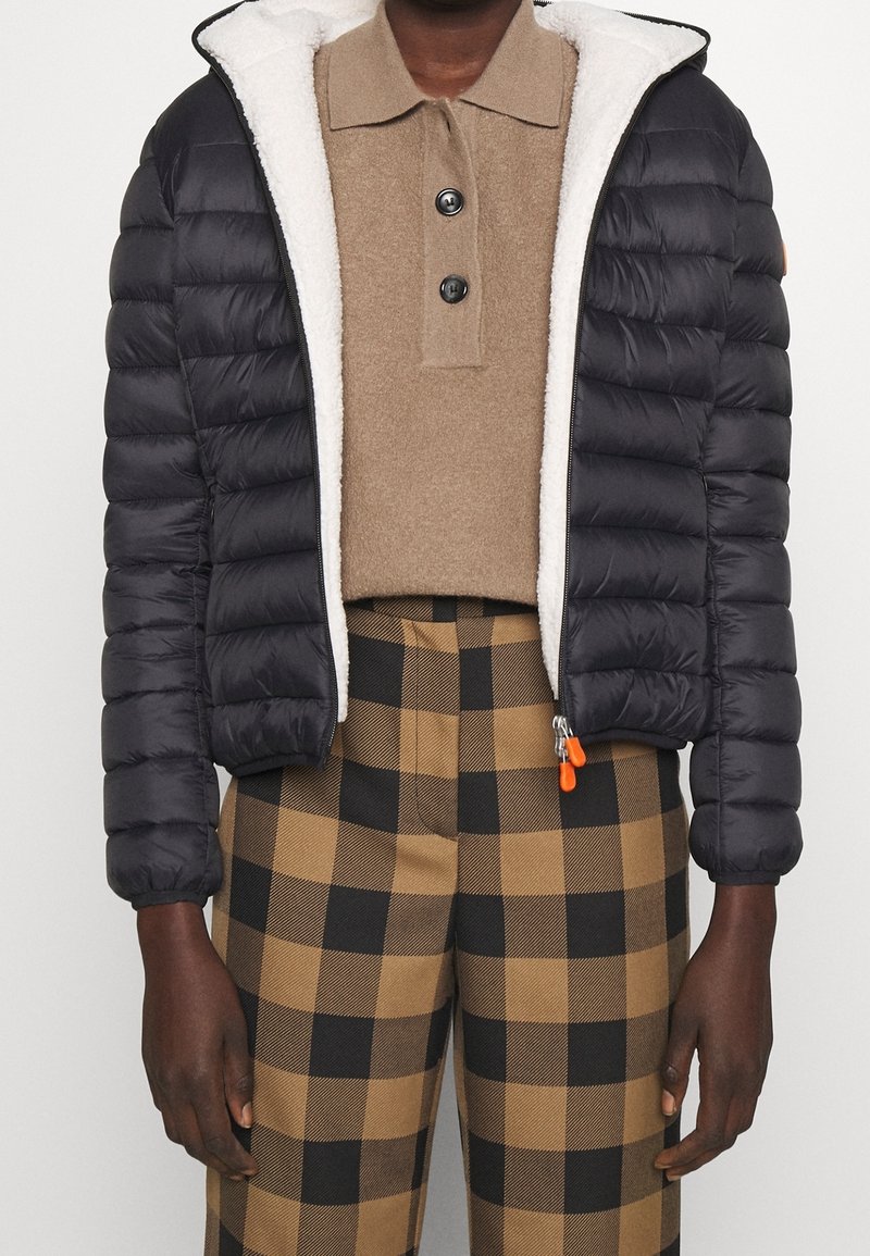 Doudoune noire avec un extérieur texturé, une capuche doublée en polaire et une fermeture éclair. Portée sur un polo beige et un pantalon à carreaux.