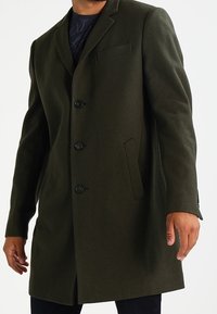 Manteau en laine vert olive avec un design à simple boutonnage, trois boutons, poches latérales et une poche poitrine. Coupe ajustée avec épaules structurées.