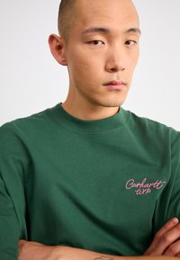 Camiseta de algodón verde con cuello redondo, que presenta un logotipo bordado en rosa "Carhartt WIP" en el lado izquierdo del pecho. Textura suave y corte relajado.