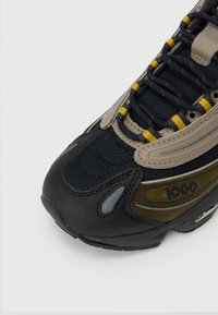 Αθλητικό παπούτσι με μαύρο επάνω μέρος από διχτυωτό υλικό, μπεζ και ελιά λεπτομέρειες, κίτρινα κορδόνια που ξεχωρίζουν και υφασμένο μαύρο καουτσούκ σόλα· διαθέτει το σήμα "1000".