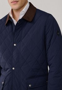 Marineblau gesteppte Jacke mit braunem Cordkragen, Druckknöpfen und Seitentaschen. Verfügt über eine rautenförmige Steppung.
