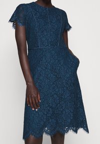 Marinblå spetsklänning med vågig nederkant, korta fransade ärmar och en figurnära design som har dekorativ sömnad och fickor.