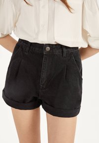 Pantalones cortos de mezclilla negros con dobladillo doblado, que cuentan con bolsillos laterales y cierre de botón en la parte delantera, combinados con una blusa blanca.