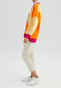 Dziergany kardigan w żółtym kolorze z pomarańczowymi i różowymi akcentami, o luźnym kroju. Noszony z beżowymi spodniami i białymi trampkami.