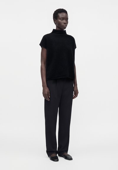 Femme portant un haut noir à col roulé à manches courtes, un pantalon noir à jambes larges et des ballerines noires, debout devant un fond clair uni.