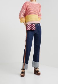 Frau in einem gestreiften rosa und gelben Pullover, dunkelhimmelblauen Jeans mit seitlichen Streifen und Bündchen, Plateausandalen und einer kleinen roten Umhängetasche mit Muster.