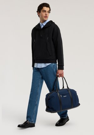 Hombre con sudadera con capucha negra, camisa azul claro, vaqueros azules y zapatos negros caminando mientras lleva una bolsa de deporte azul marino en la mano derecha.