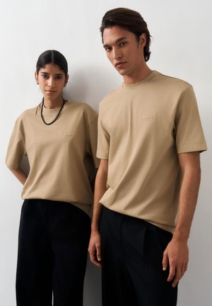 Deux mannequins portant des T-shirts beige BOSS et des pantalons noirs se tiennent devant un fond blanc uni, regardant droit devant eux avec des expressions neutres.