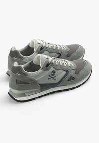 Graue Turnschuhe mit einem Design aus Wildleder und Mesh, ausgestattet mit einem Totenkopf- und Querbones-Logo, dicker Gummisohle und kontrastierenden blauen Akzenten.