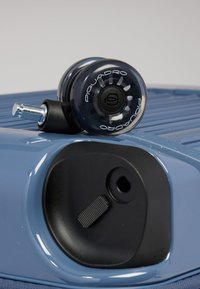 Gros plan sur un coin d'une valise bleue avec un mécanisme de verrouillage noir et une roulette pivotante transparente portant la marque "Piquadro".