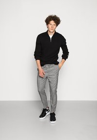Sweater preto com fecho, calças cinza com padrão xadrez e ténis pretos com solas brancas. Modelo em pé com uma postura relaxada contra um fundo simples.