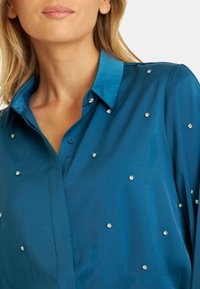Blusa de satén en color verdoso con cuello clásico, cierre de botones y piedras embellecedoras dispersas para mayor textura y detalle.