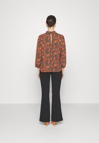 Blusa a maniche lunghe floreale in marrone scuro, con motivi arancioni e verdi, con scollatura arricciata e chiusura a chiave sul retro. Abbinata a pantaloni svasati neri.