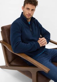 Pullover in pile blu navy con zip a quarto, dal tessuto morbido e logo discreto, indossato sopra una camicia blu, abbinato a jeans scuri, seduto su una sedia di legno.