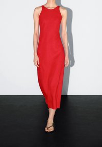 Femme portant une robe midi rouge sans manches et des sandales marron, marchant sur un sol noir avec un fond blanc uni.