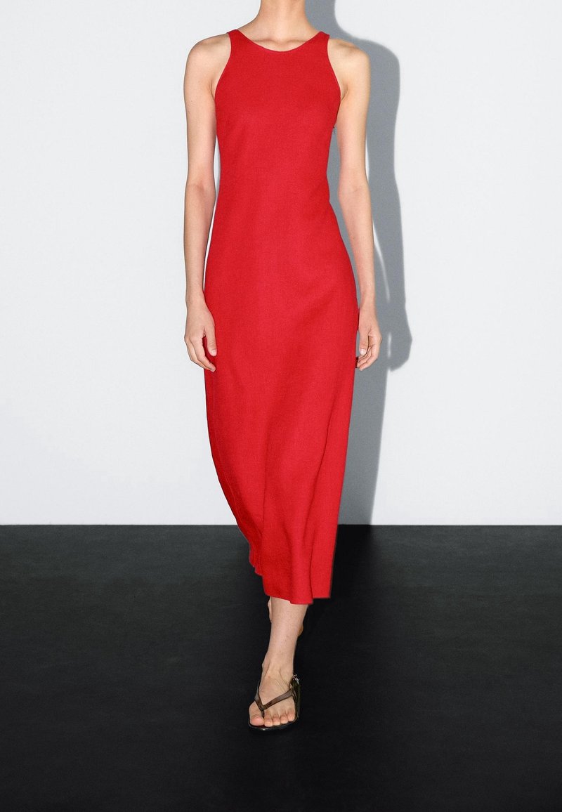 Femme portant une robe midi rouge sans manches et des sandales marron, marchant sur un sol noir avec un fond blanc uni.