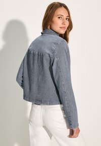 Chaqueta de mezclilla a rayas con un diseño recortado, que presenta cuello y mangas largas. Se muestra con pantalones blancos, destacando un contraste de colores.