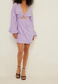Lila Kleid mit tiefem V-Ausschnitt, gedrehtem Vorderteil und langen, luftigen Ärmeln. Aus strukturiertem Stoff; oberschenkellang. Braune hochhackige Sandalen.