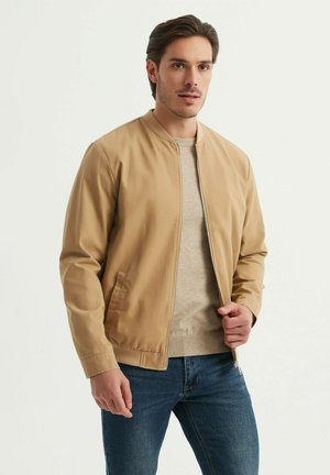Veste bomber beige en tissu lisse, avec fermeture éclair à l'avant, col côtelé et poches latérales, portée par-dessus un pull léger et un jean.