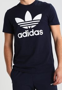 Námořnická modrá bavlněná triko s velkým bílým logem Adidas s klasickým trojlístkovým designem. Kulatý výstřih a krátké rukávy.