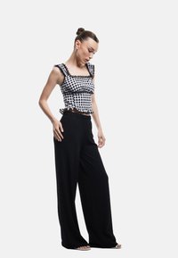 Zwart-witte gingham crop top met gerende schouderbandjes en zoom, gecombineerd met loszittende zwarte broek. Model staat in een neutrale setting.