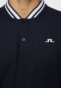 Marinblå polotröja med en texturerad tyg. Har en randig krage i vitt och marinblått, en treknappsstängning och en vit logotyp på bröstet.