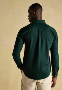 Mann in einem dunkelgrünen Langarmshirt mit dezenten vertikalen Streifen, beigen Hosen, steht vor einem schlichten hellen Hintergrund, von hinten betrachtet.