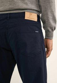Chinos azul marino de algodón con una etiqueta de cuero en la parte trasera de la cinturilla que presenta el logotipo de la marca y un detalle en el bolsillo trasero.
