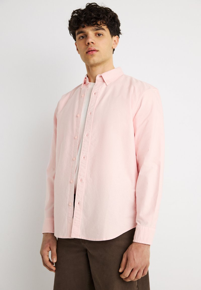 Jeune homme aux cheveux bouclés portant une chemise rose clair boutonnée sur un t-shirt blanc et un pantalon marron foncé, debout devant un fond blanc.
