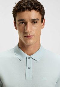 Light blue polo shirt with a textured fabric, two-button placket, and a subtle embroidered logo on the left chest.